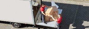 Deux déménageurs en chemise rouge chargent des cartons dans un camion de déménagement blanc, l'un à l'intérieur, l'autre à l'extérieur, sur un pavé. Travail d'équipe lors du déménagement.