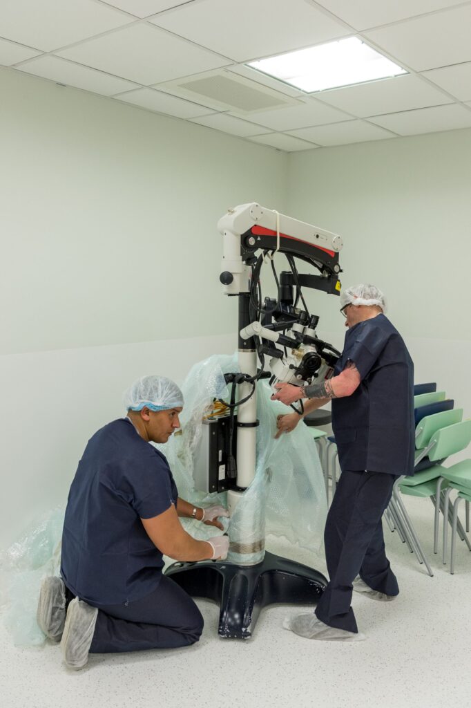 Deux professionnels en tenue médicale, charlotte et sur-chaussures, installent un grand microscope chirurgical dans une salle d'opération lumineuse.