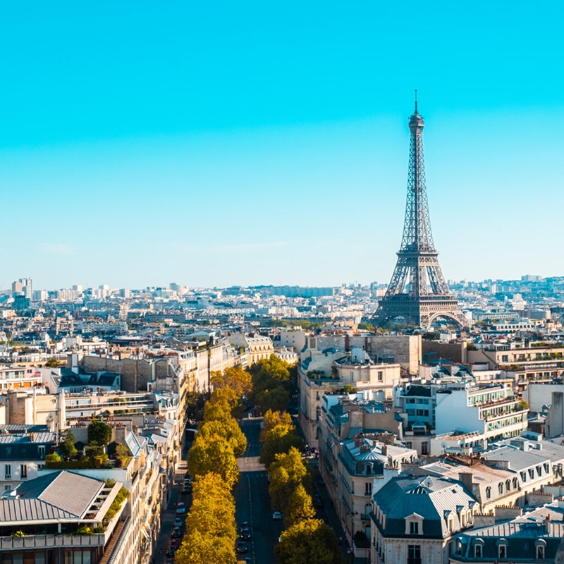 Vue aérienne de Paris sous ciel bleu, la Tour Eiffel dominant les toits haussmanniens et une rue bordée d'arbres dorés.