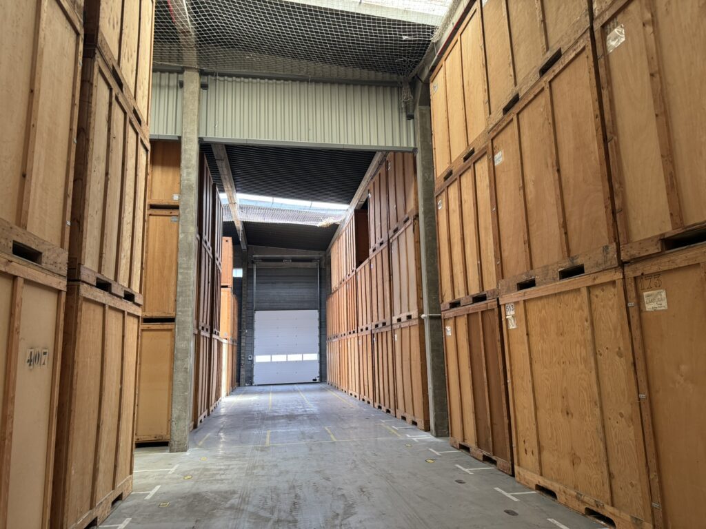 Un couloir d'entrepôt bordé de grandes caisses de rangement en bois, numérotées 407, 173, 513, et avec l'inscription 'ZANDUREAU'. Une porte de garage blanche est visible au fond.