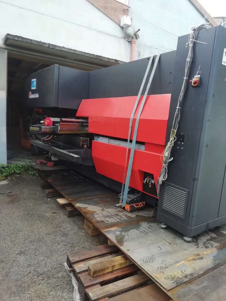 Grande machine industrielle AMADA (rouge/noir) sur palettes, sanglée pour déménagement/installation dans un entrepôt.