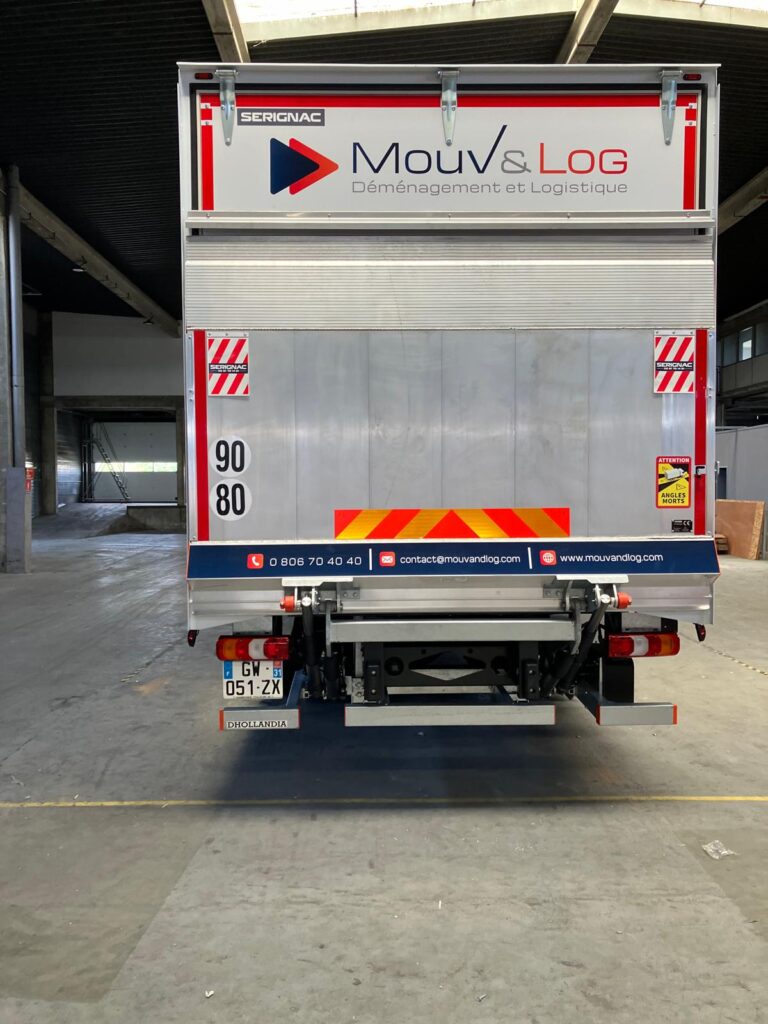 Arrière d'un camion de déménagement Mouv&Log en entrepôt, affichant contacts (tél, mail, web), vitesses 90/80, et avertissement angles morts.
