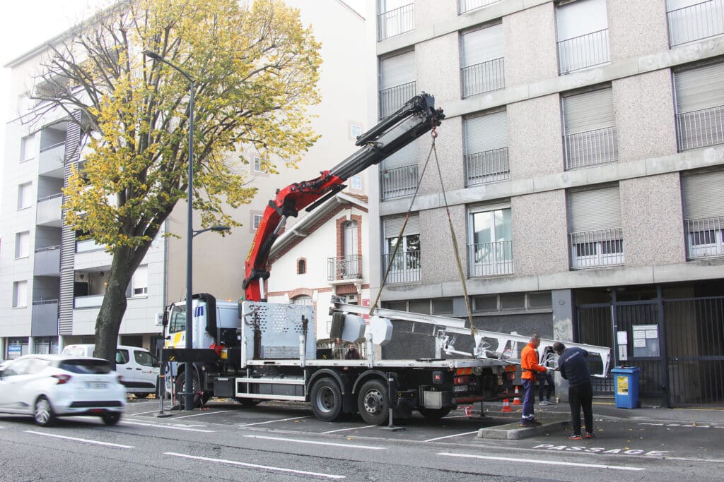 Camion-grue rouge déchargeant de longs panneaux blancs devant un immeuble gris et un arbre aux feuilles jaunes. Deux ouvriers manipulent les matériaux au sol. Texte visible : PASSAGE.