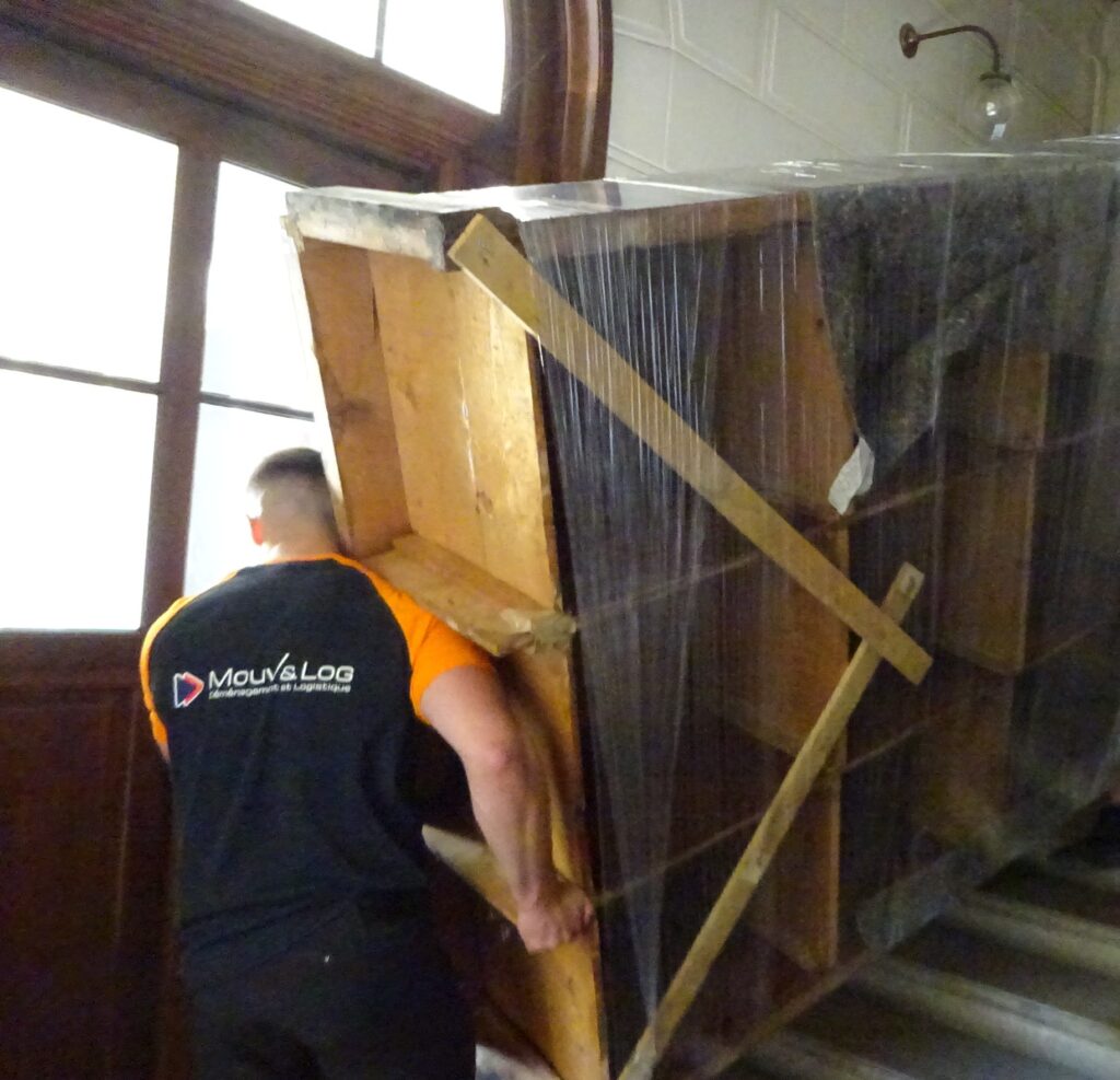 Déménageur Mouv&Log (Déménagement et Logistique) monte un meuble lourd et emballé, sécurisé par du bois, dans un escalier.