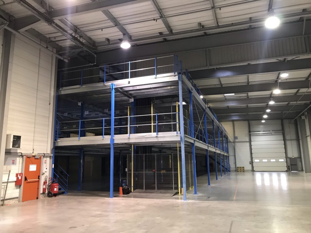 Entrepôt logistique vide, mezzanine industrielle à deux niveaux, porte d'urgence orange et extincteur. Grande porte de quai, plafond éclairé.