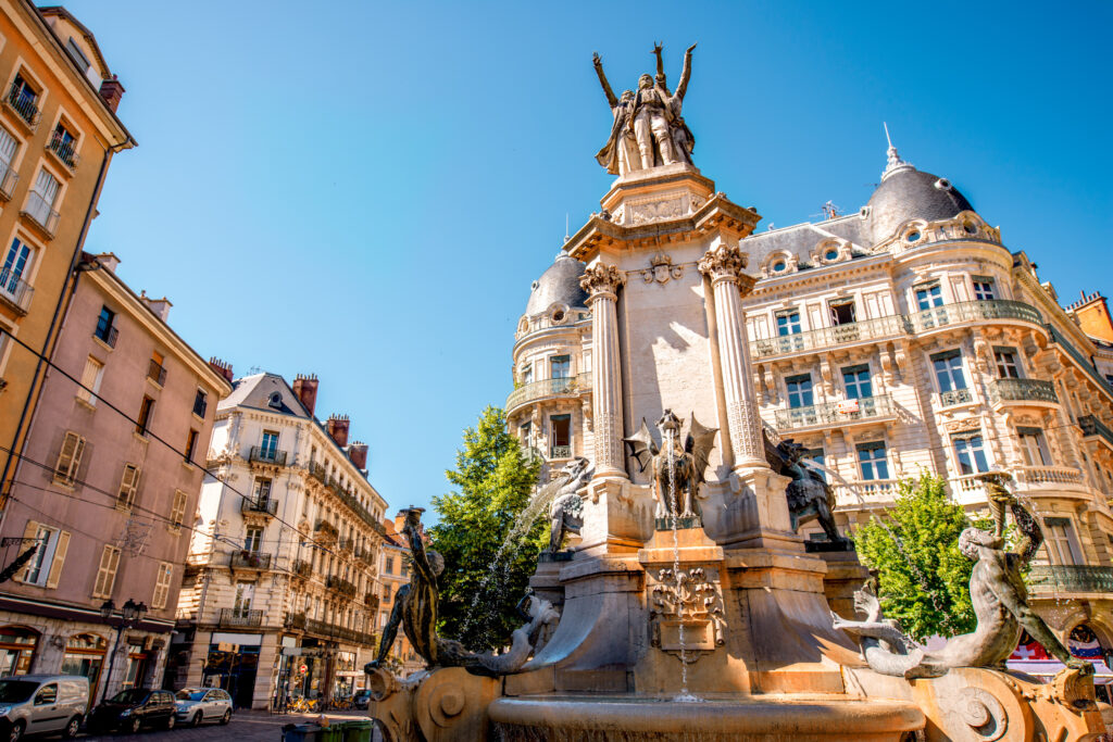 Grenoble: Fontaine des Trois Ordres, Place Victor Hugo Fontaine des Trois Ordres de Grenoble avec statues aux bras levés, mermen et gargouilles. Bâtiments classiques sous ciel bleu clair.