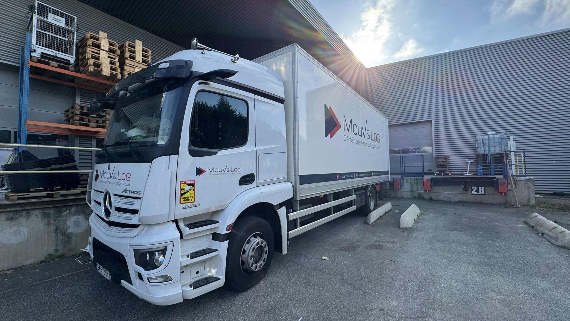 Camion Mercedes-Benz Actros MouV&Log blanc devant entrepôt. Déménagement et Logistique, 'ATTENTION ANGLES MORTS'. Quai de chargement '2B'.