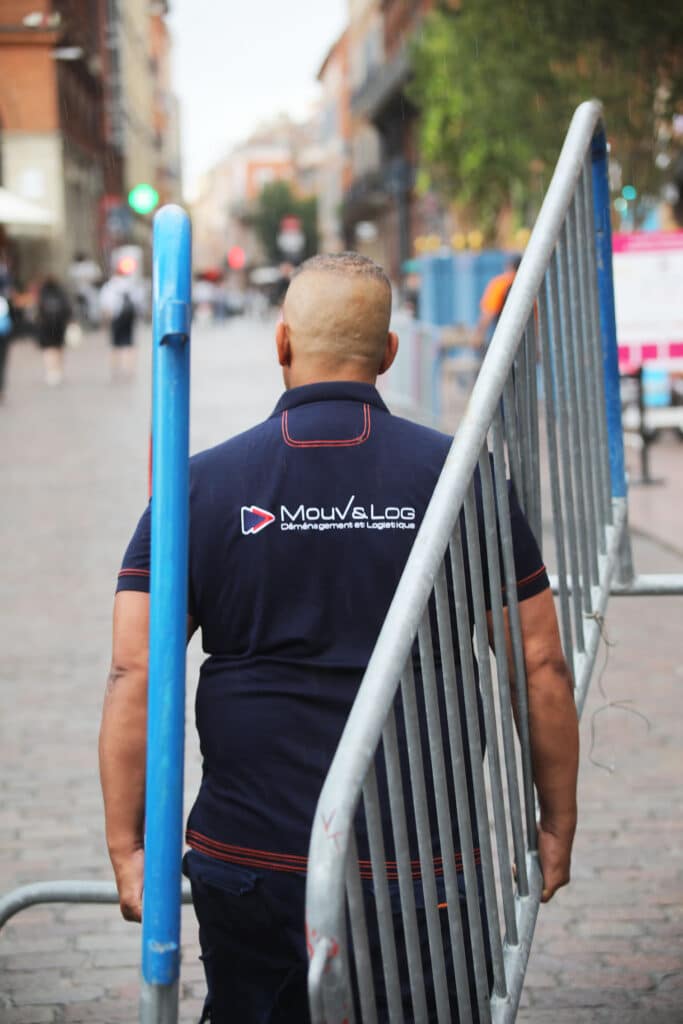 Un homme de dos, en polo MouV&Log Déménagement et Logistique bleu marine, déplace une barrière métallique sous la pluie.