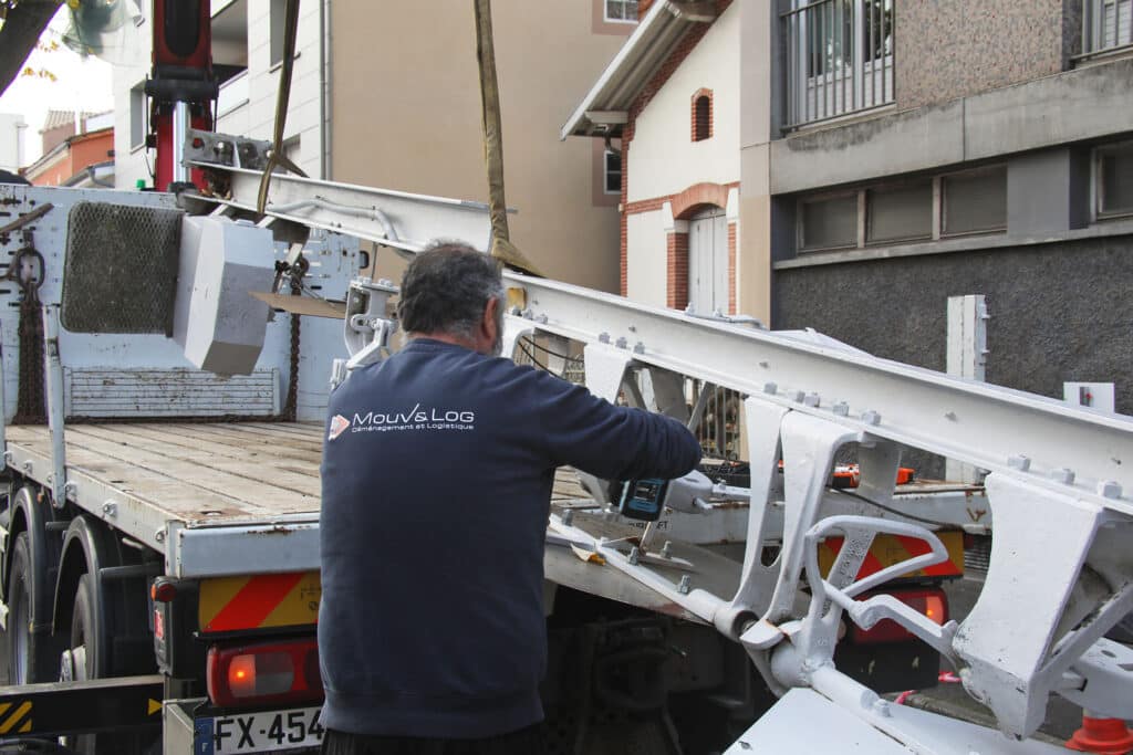 Un employé Mouv's Log avec le camion FX-454 assemble des structures métalliques blanches à l'aide d'un outil électrique, sous une grue de levage.