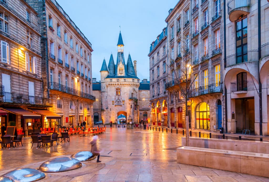 Porte Cailhau Bordeaux : Soirée historique et lumière Porte Cailhau, Bordeaux, au crépuscule. La porte médiévale et les façades éclairées se reflètent sur les pavés mouillés. Une personne marche.
