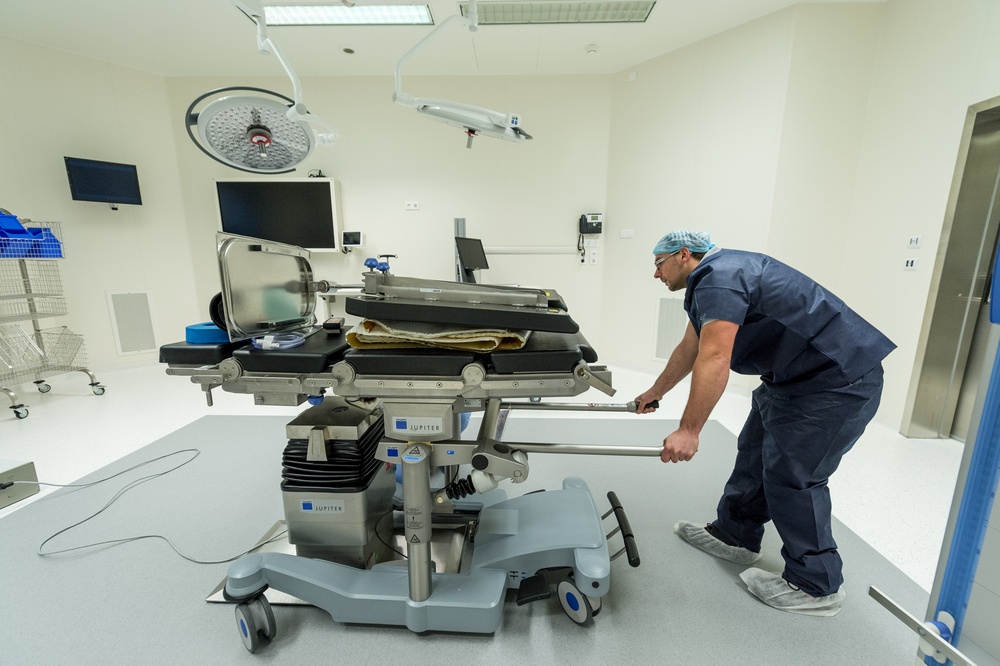 Professionnel de santé pousse table d'opération JUPITER dans un bloc chirurgical moderne. Salle lumineuse avec écrans, éclairages médicaux, équipement.