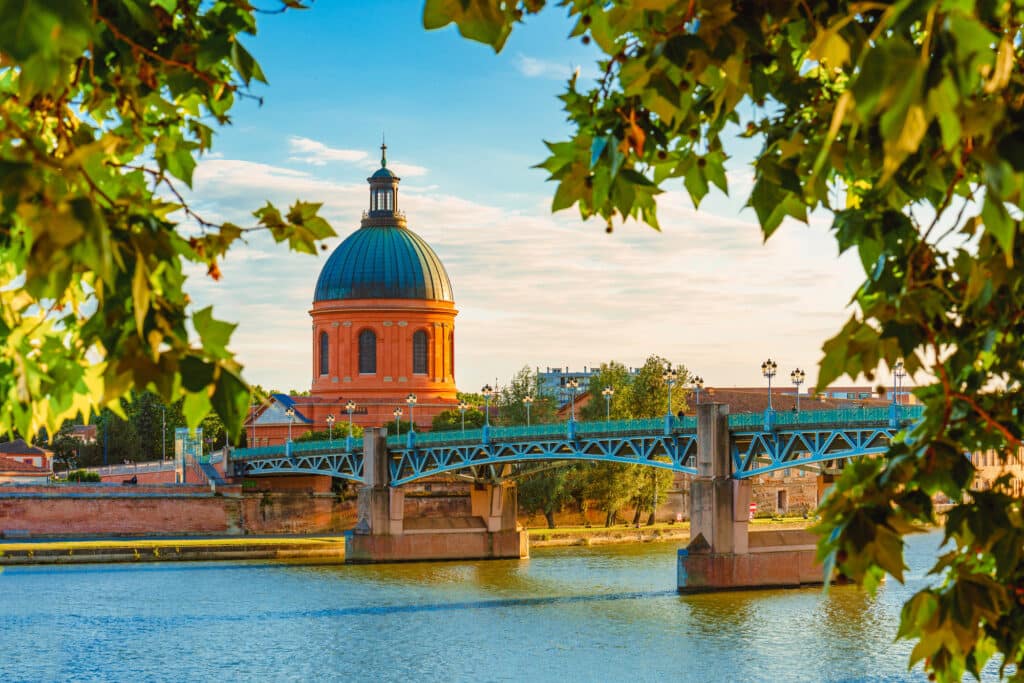 Toulouse: Dôme de la Grave, Pont Saint-Pierre sur la Garonne Dôme de la Grave et Pont Saint-Pierre enjambant la Garonne à Toulouse, sous un ciel clair, encadrés de feuillage vert.