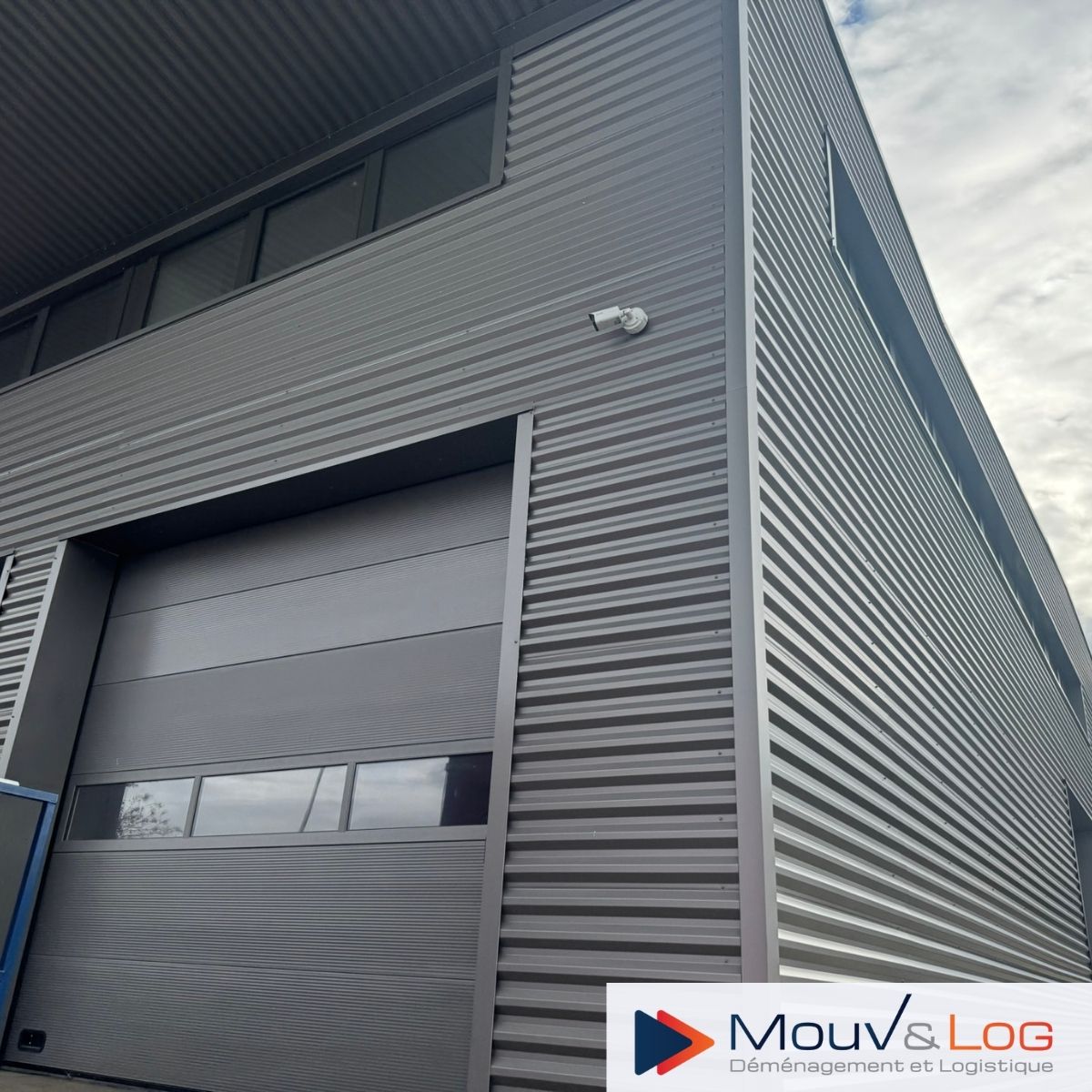 Bâtiment Mouv&Log Déménagement et Logistique, entrepôt industriel gris avec porte de garage et sécurité.
